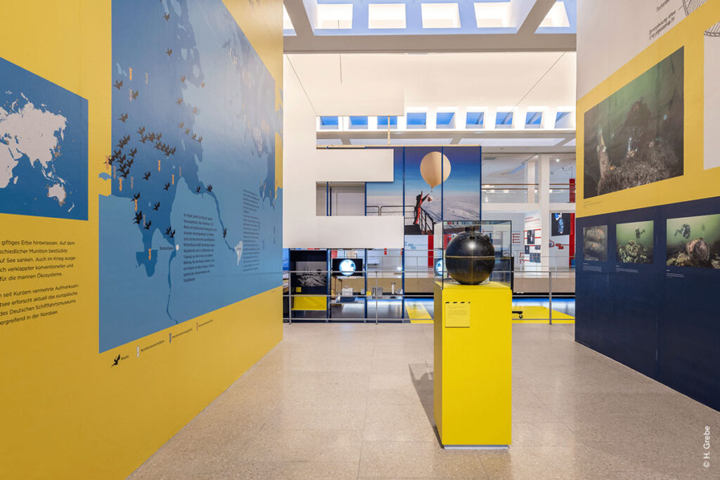 Museumsraum der Ausstellung „CHANGE NOW“ mit Vitrinen-Exponat, Fotografien und Wandgrafiken zu Schiffswracks.